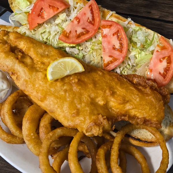 Golden-fried fish sandwich with lettuce, tomato, and a lemon wedge, served with onion rings.