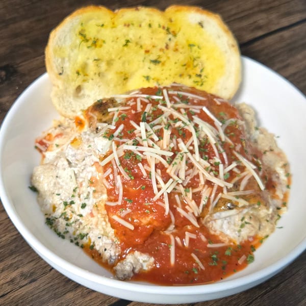 A white bowl of baked ziti with red sauce, cheese, and parsley, served with garlic bread.