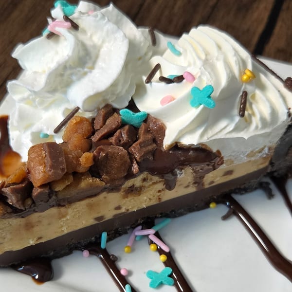 A slice of peanut butter pie with whipped cream, chocolate drizzle, and colorful sprinkles.