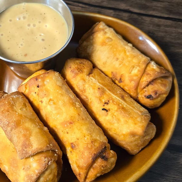 Four crispy egg rolls on a brown plate with a side of creamy dipping sauce.
