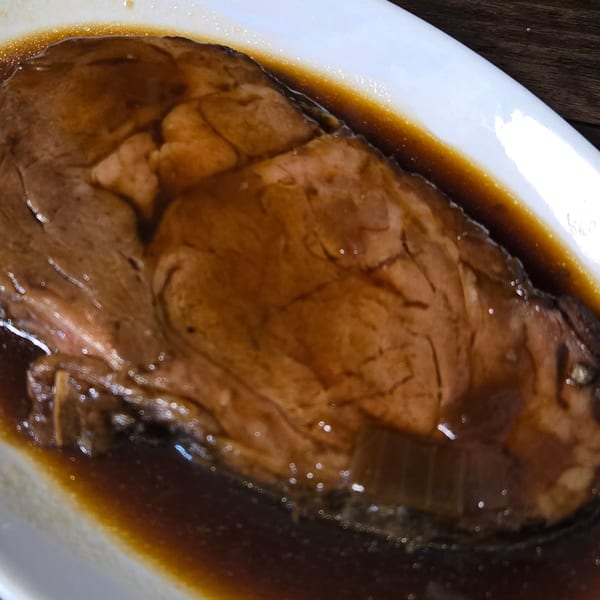 A large, glistening slice of roast beef swimming in a rich, dark brown gravy on a white plate.